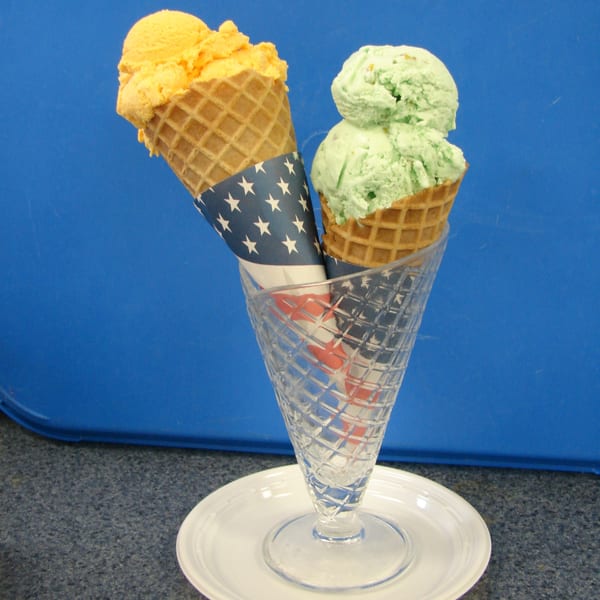 Two ice cream cones, one orange and one green, in a glass holder against a blue background.