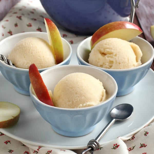 Three light-colored ice cream scoops in blue bowls, garnished with red apple slices, on a white plate.