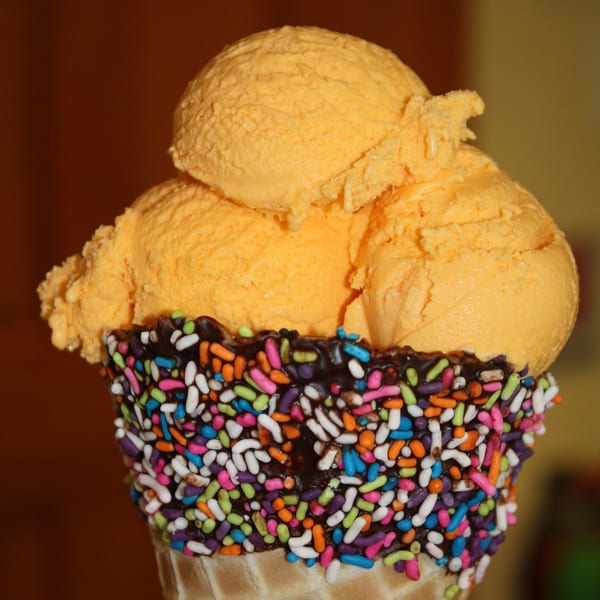 Three scoops of orange ice cream in a waffle cone with chocolate and colorful sprinkles.