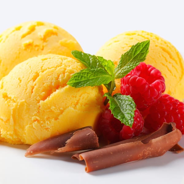 Three scoops of yellow ice cream with raspberries, mint, and chocolate curls on a white background.